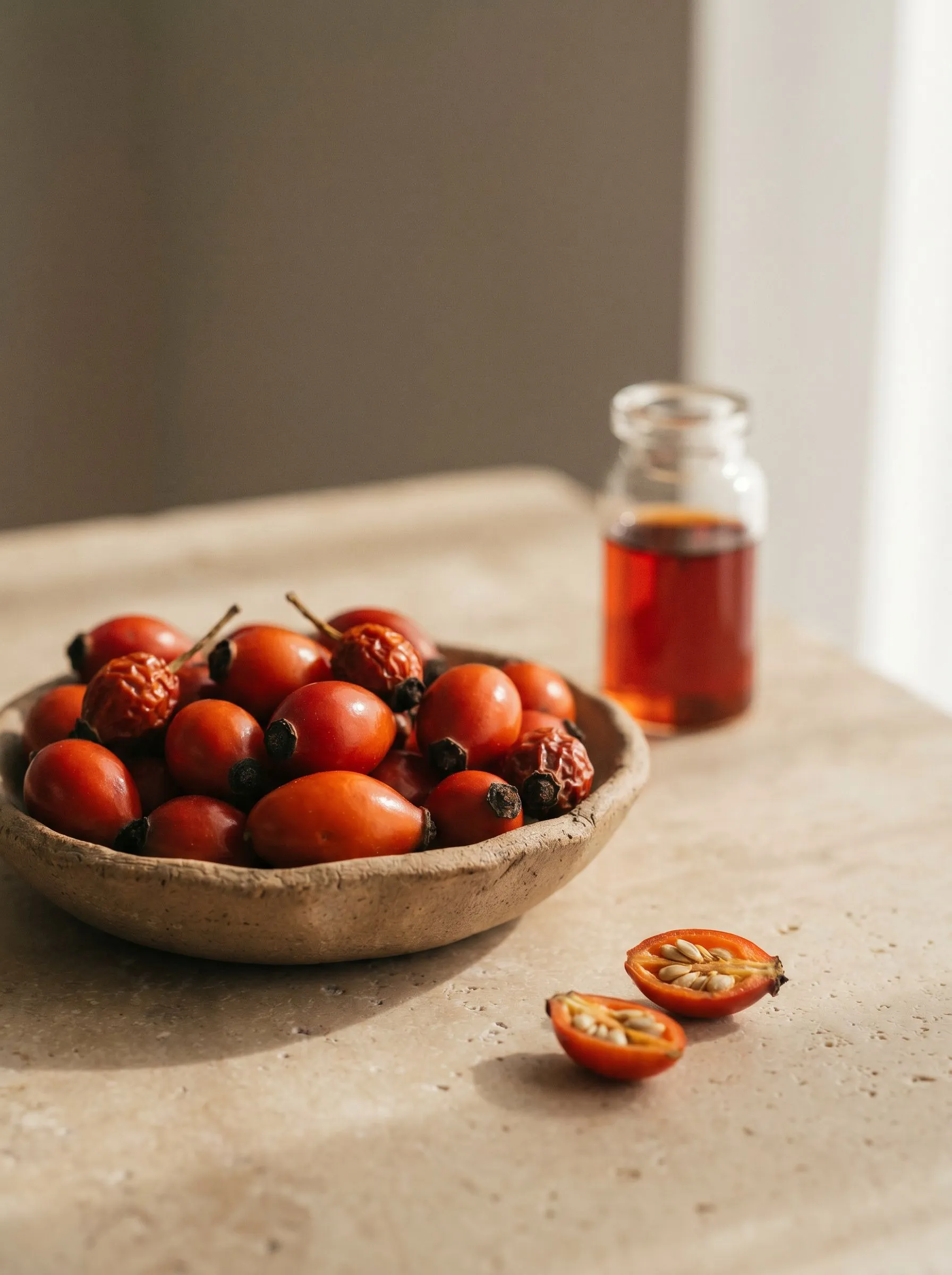 Fresh rosehips in a clay dish beside a vial of rosehip oil